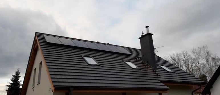 Czarny dach z dachówką ceramiczną, na którym zamontowano panele fotowoltaiczne i okna dachowe, widok na komin z blachy na tle pochmurnego nieba.