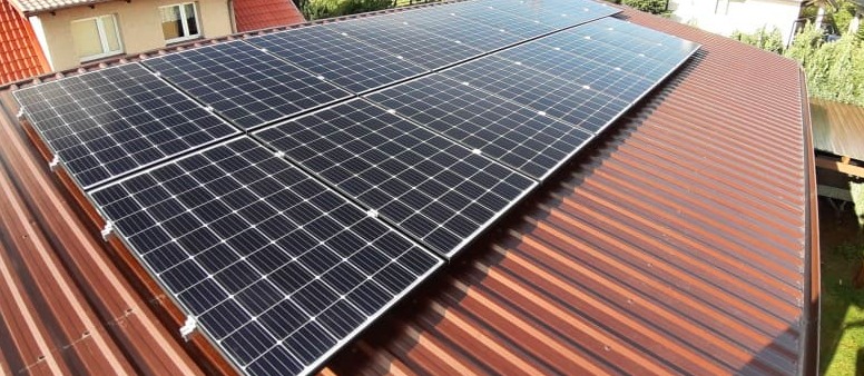 Panele fotowoltaiczne zamontowane na metalowym dachu z widocznymi mocowaniami, z fragmentem budynku w tle.