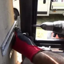 Montaż okna: wiertarka w dłoniach w rękawicach przykręca metalowy element do ramy okiennej, w tle biały samochód dostawczy.