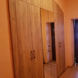 Wysoka szafa z drewnopodobnymi frontami i lustrzanymi drzwiami, zwieńczona łukowym elementem, w pomieszczeniu o pomarańczowych ścianach.