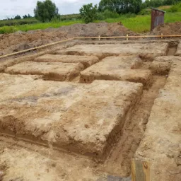 Wykopane podłoże pod fundamenty z drewnianym szalunkiem, widoczne przygotowane rowy i hałdy ziemi w tle.