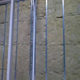 Montaż izolacji z wełny mineralnej pomiędzy metalowymi profilami na ścianie wewnętrznej budynku, widoczne elementy konstrukcyjne sufitu.