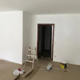 Wnętrze pokoju po odświeżeniu ścian na biało, widoczna drabina malarska, wiadro z farbą oraz sprzęt malarski na zabezpieczonej podłodze, ciemna ościeżnica drzwi.