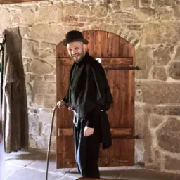 Uśmiechnięty mężczyzna w czarnym stroju i meloniku, oparty o laskę, stoi na tle kamiennej ściany i drewnianych drzwi.