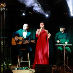 Zesp&oacute;ł muzyczny na scenie: wokalistka w czerwonej sukni śpiewa do mikrofonu, gitarzysta w zielonym garniturze gra na gitarze akustycznej, a perkusista gra na elektronicznej perkusji. Scena...