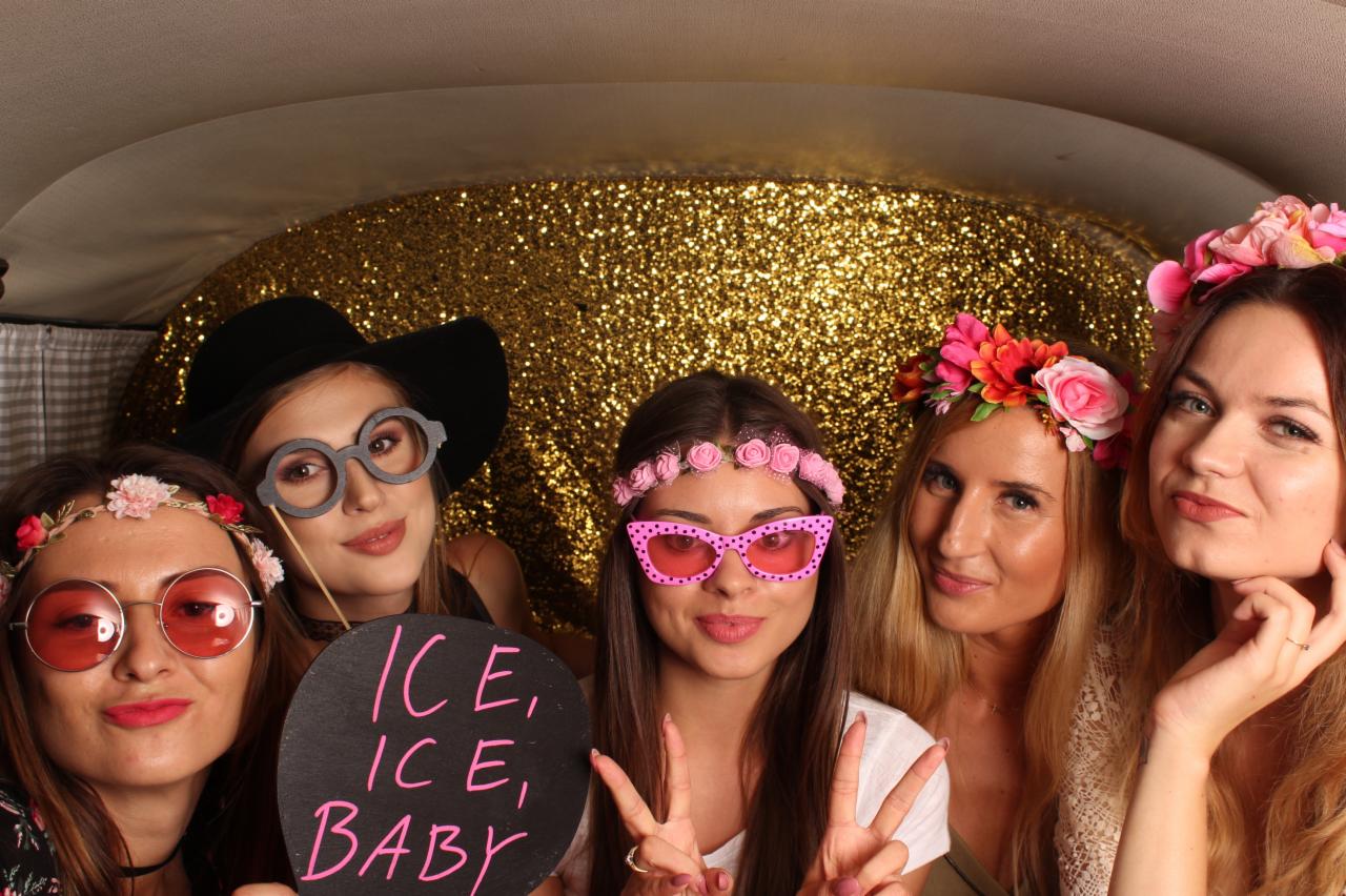 Grupa pięciu uśmiechniętych kobiet pozuje w fotobudce z brokatowym tłem, używając rekwizytów takich jak okulary, kapelusz i tabliczka z napisem 'Ice, Ice, Baby'.