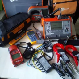 Zestaw różnorodnych urządzeń i akcesoriów pomiarowych, w tym mierniki marki Sonel, Hilti i Fluke, kable, cęgi prądowe i torby transportowe, rozłożonych na jasnym blacie.