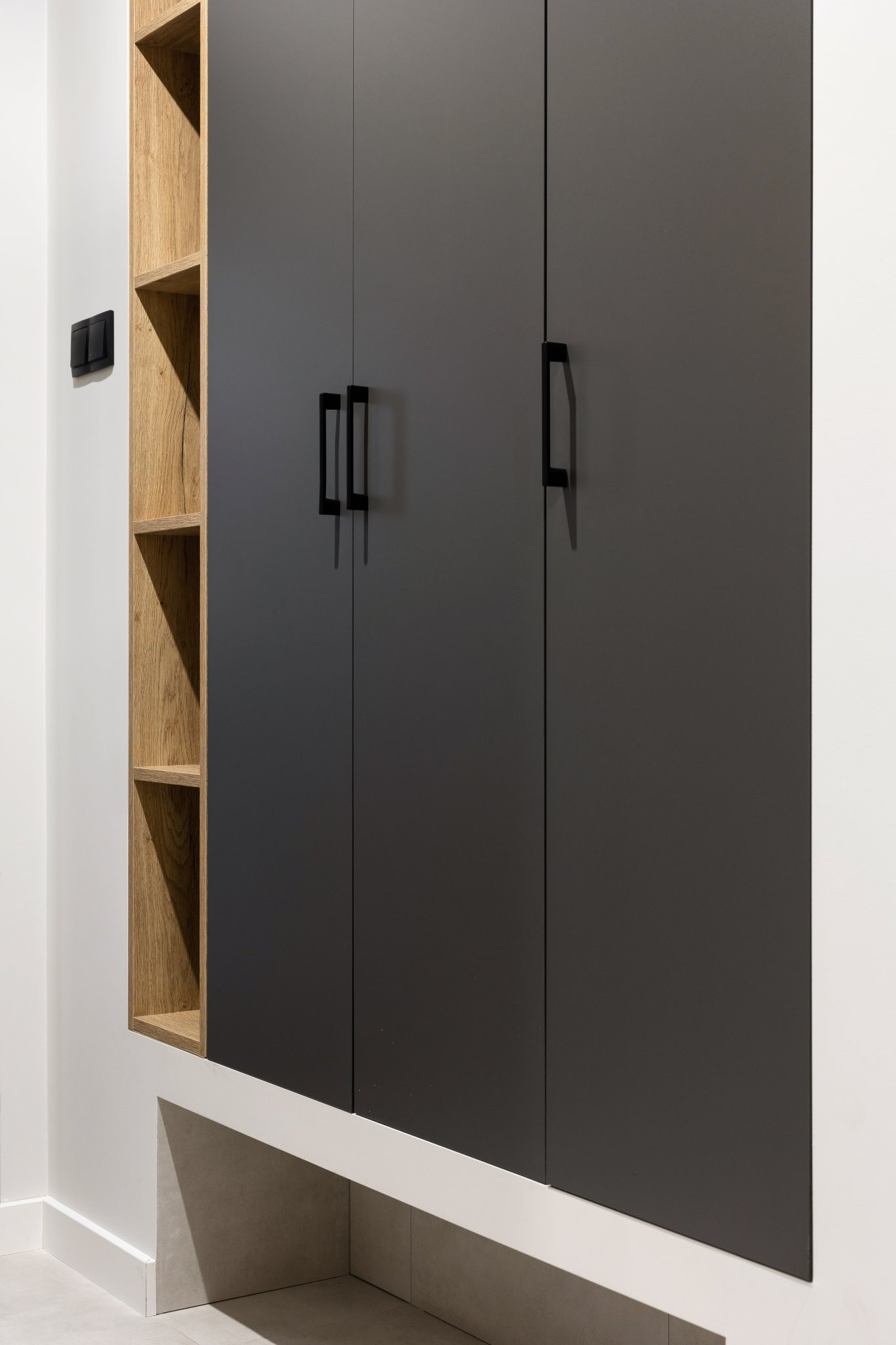 Ciemnoszare szafki z czarnymi uchwytami, z otwartymi drewnianymi półkami po lewej stronie, na tle białej ściany z czarnym włącznikiem światła.