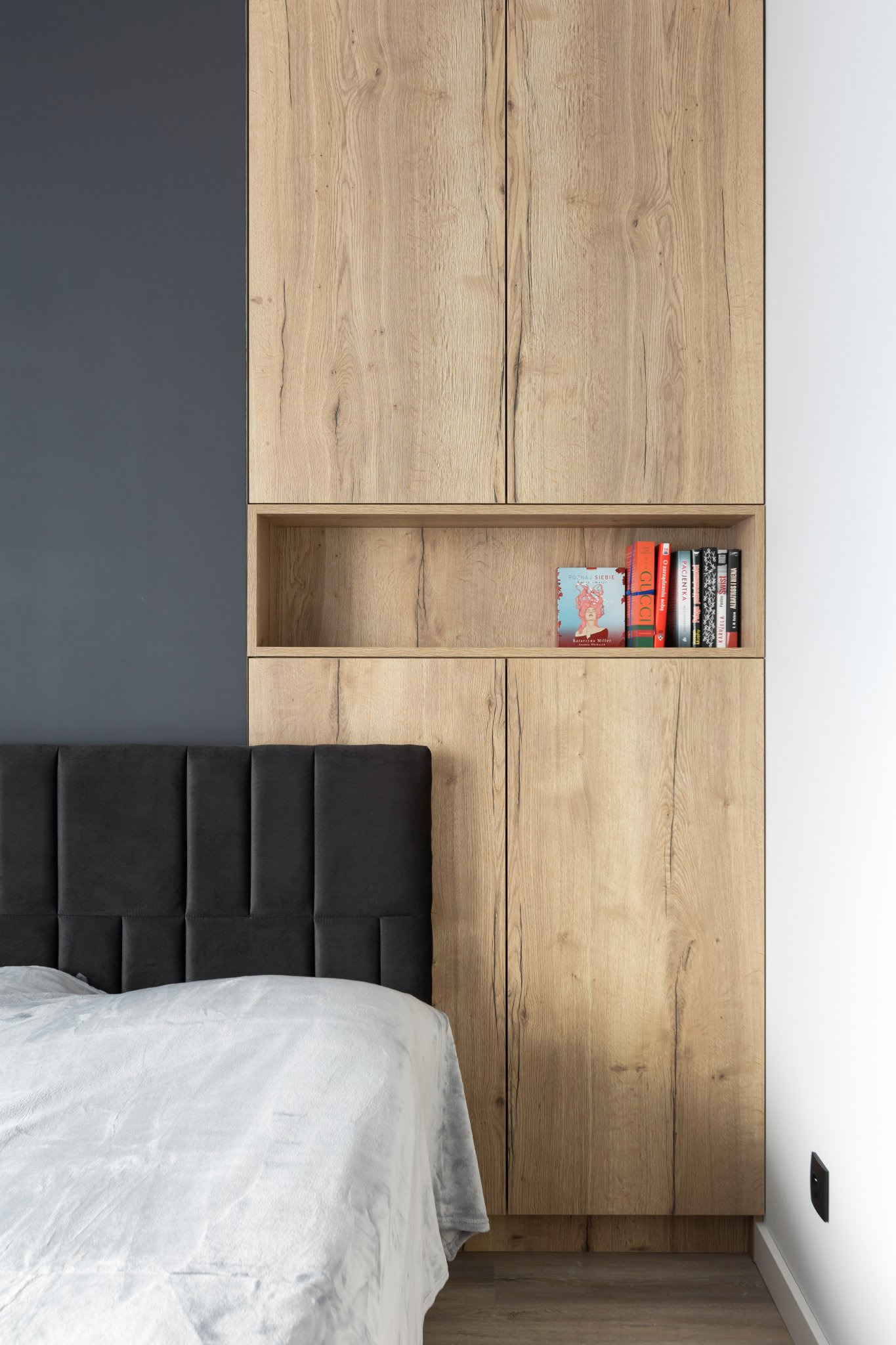 Drewniana szafa wnękowa z półką na książki nad łóżkiem z szarym wezgłowiem i białą pościelą, widok z bliska na szafę i fragment łóżka.