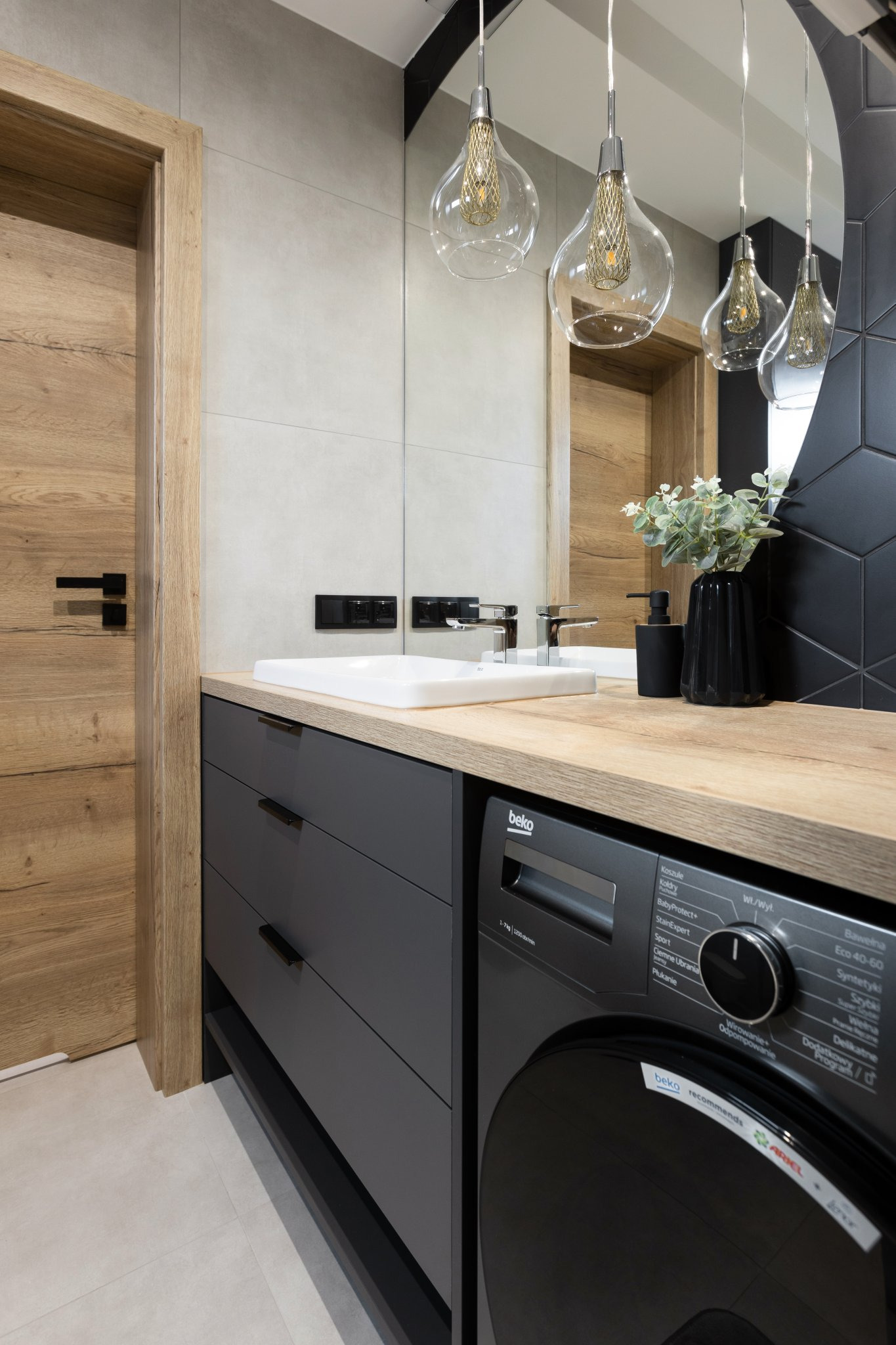 Nowoczesna łazienka z drewnianymi drzwiami, szarymi meblami podumywalkowymi, pralką Beko i dekoracyjnymi lampami wiszącymi.