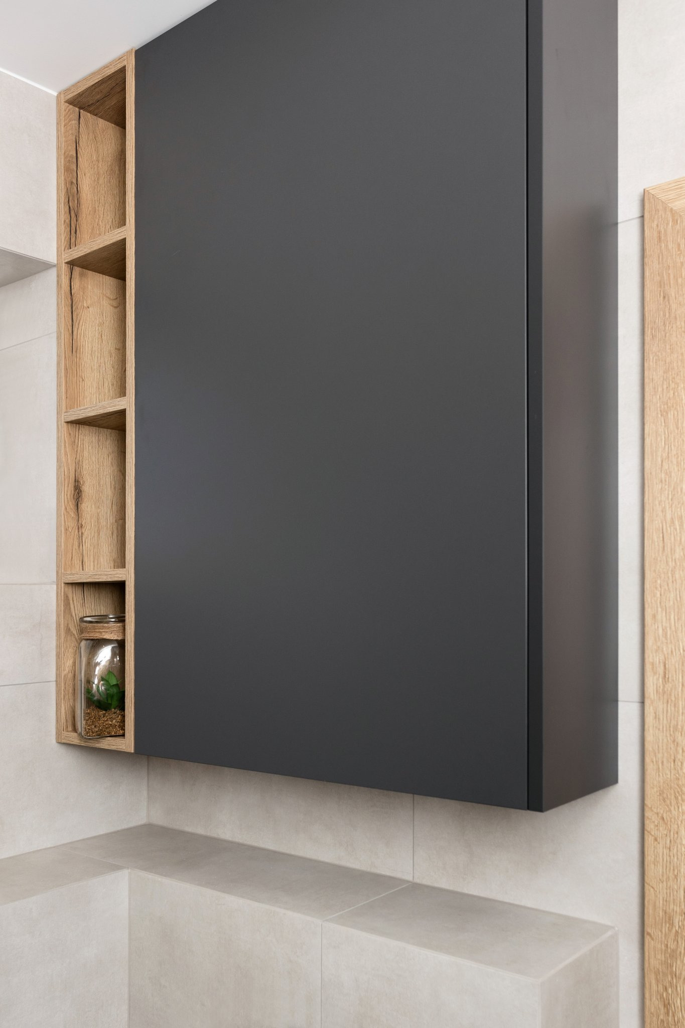 Szara szafka wisząca z drewnianymi półkami po lewej stronie, na jednej z nich słoik z rośliną.