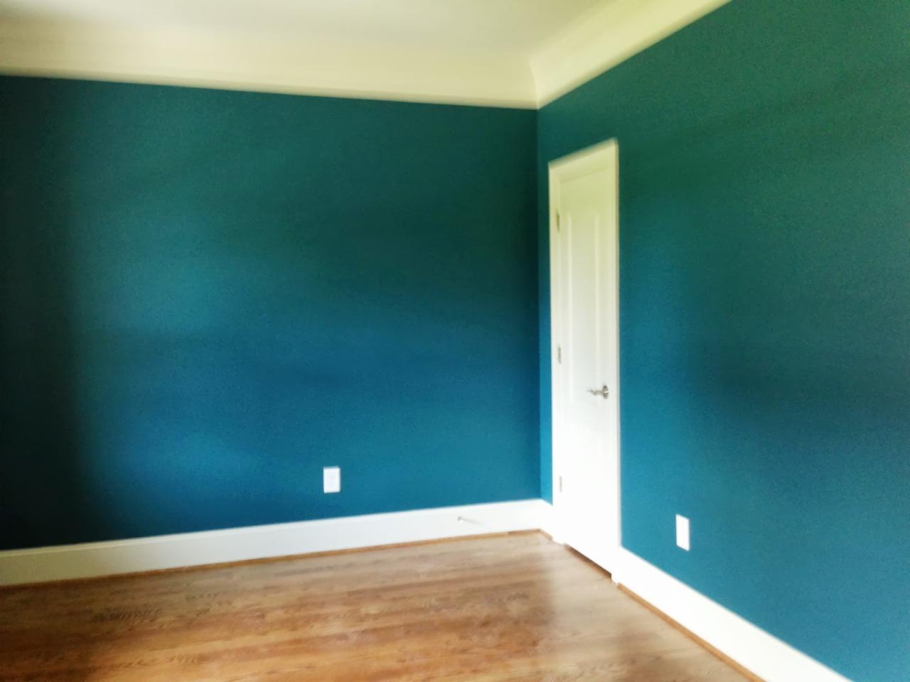 Pomalowane na turkusowo ściany w pustym pokoju z drewnianą podłogą i białymi listwami przypodłogowymi oraz białymi drzwiami.