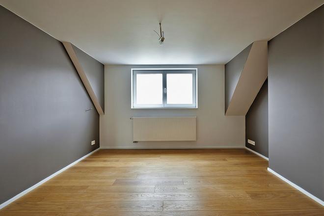 Puste poddasze z drewnianą podłogą, świeżo pomalowane ściany w odcieniach szarości i bieli, okno z białymi ramami i grzejnik pod nim, surowe oświetlenie sufitowe.