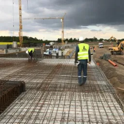 Zbrojenie fundamentów pod budowę hali, widoczne dwie pracujące osoby w odblaskowych kamizelkach, żurawie budowlane w tle i pochmurne niebo.