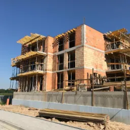 Wznoszenie trzypiętrowego budynku z czerwonej cegły, widoczne rusztowania i szalunki, fundamenty z izolacją termiczną, błękitne niebo w tle.