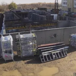 Budowa fundamentów domu wielorodzinnego z widocznymi szalunkami, zbrojeniami i sprzętem budowlanym na placu budowy w słoneczny dzień.