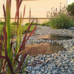 Ogród z ścieżką z kamiennych płyt, żwirem, ozdobną trawą o czerwono-zielonych liściach oraz kulistą rośliną i lawendą w tle.