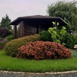 Starannie zaprojektowany ogród z drewnianą altaną, kolorowymi krzewami i równo przystrzyżonym trawnikiem w pochmurny dzień.