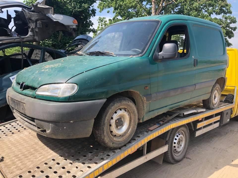 Zielony, lekko zabrudzony Peugeot Partner na lawecie, w tle wrakowane pojazdy na złomowisku.