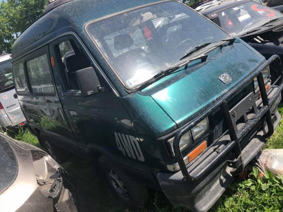 Zielony, lekko zakurzony van Subaru 4WD z czarnym orurowaniem z przodu, stojący na trawiastym terenie wśród innych uszkodzonych pojazdów na złomowisku.