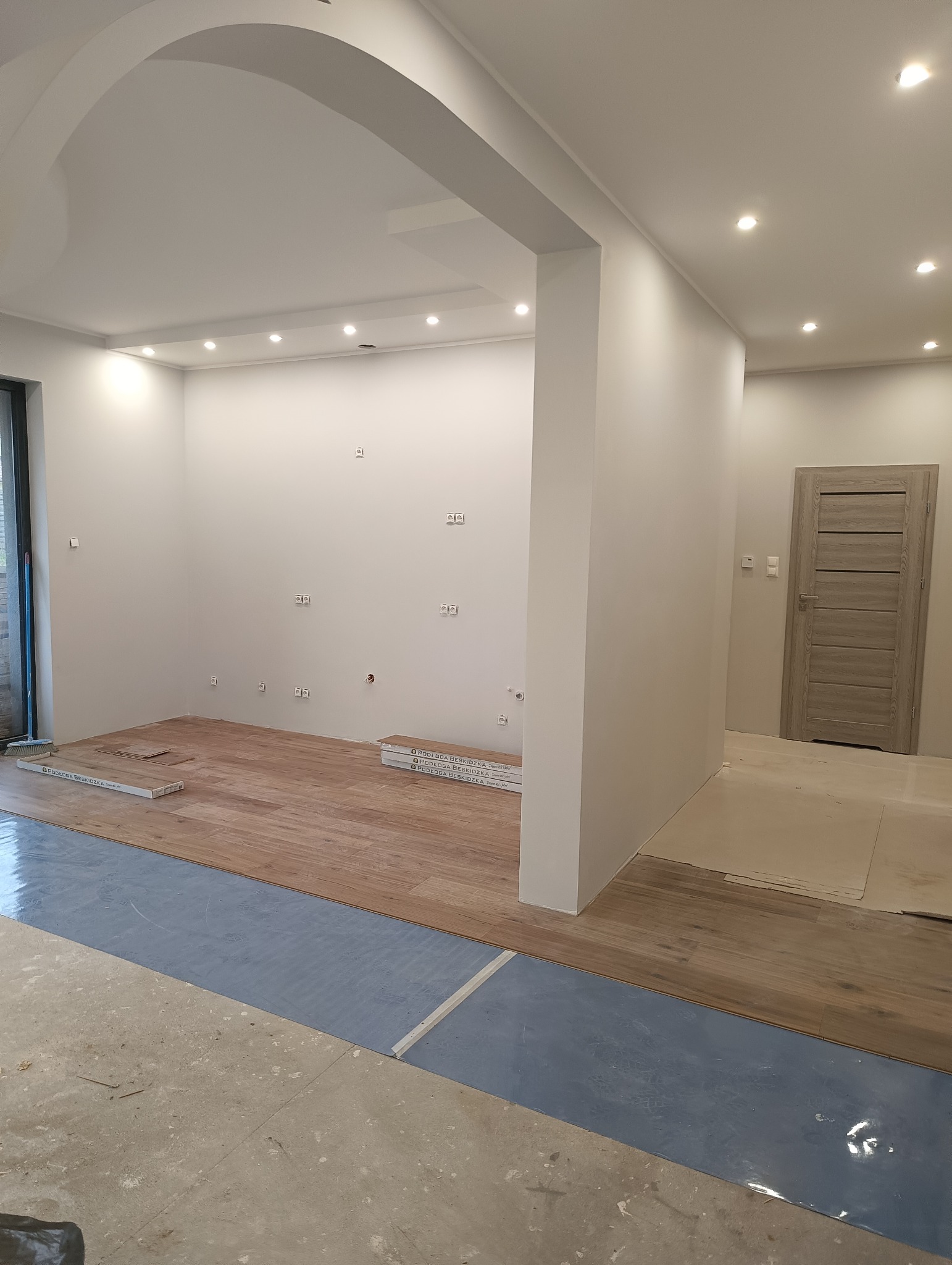 Wnętrze mieszkania w trakcie remontu: częściowo położona drewniana podłoga, zabezpieczona folią podłoga w tle, widoczne puszki elektryczne na ścianach i sufit podwieszany z oświetleniem punktowym.