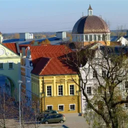 Panorama kolorowych kamienic z czerwonymi dachami, widoczna brama z dekoracjami, kopuła w tle i drzewa bez liści na pierwszym planie.