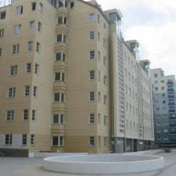 Wielopiętrowy budynek mieszkalny z beżową elewacją i szklaną nadbudówką, widoczny z poziomu placu z niskim, białym, okrągłym murem.