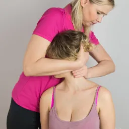 Kobieta w różowej koszulce delikatnie manipuluje szyją pacjentki siedzącej na krześle, skupiając się na odcinku szyjnym kręgosłupa.