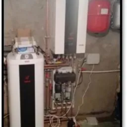 Kocioł gazowy kondensacyjny z zasobnikiem ciepłej wody, połączony miedzianymi rurami, zamontowany na tle surowej, betonowej ściany w piwnicy.