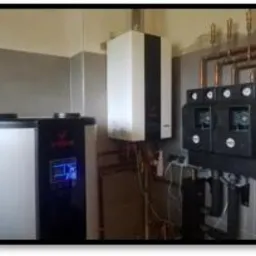 Zestaw urządzeń grzewczych w kotłowni: biała pompa ciepła z wyświetlaczem, wiszący biały piec gazowy oraz czarny rozdzielacz z manometrami, na tle wykafelkowanej ściany.