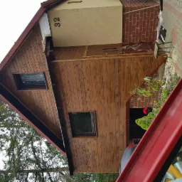 Drewniany dom z numerem 32, widoczne dwa okna i fragment dachu, z boku domu fragment chodnika z cegły i mały stołek, w tle drzewa.