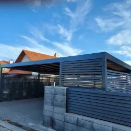 Nowoczesna, grafitowa konstrukcja carportu z poziomymi listwami, przylegająca do domu z czerwoną dachówką, widoczna zza szarego, betonowego ogrodzenia.