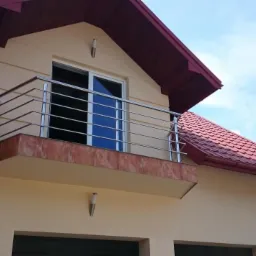 Balkon z nierdzewną balustradą, widok z dołu, jasna elewacja domu z czerwoną dach&oacute;wką i niebieskim niebem w tle.