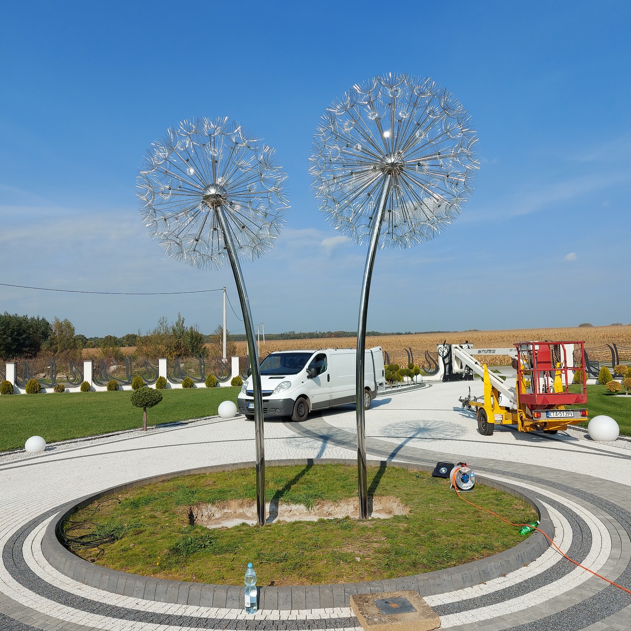 Dwa metalowe dmuchawce na trawniku, biały bus i podnośnik koszowy w tle, błękitne niebo. Dekoracja ogrodowa ze stali nierdzewnej, podświetlana od dołu, na posesji.