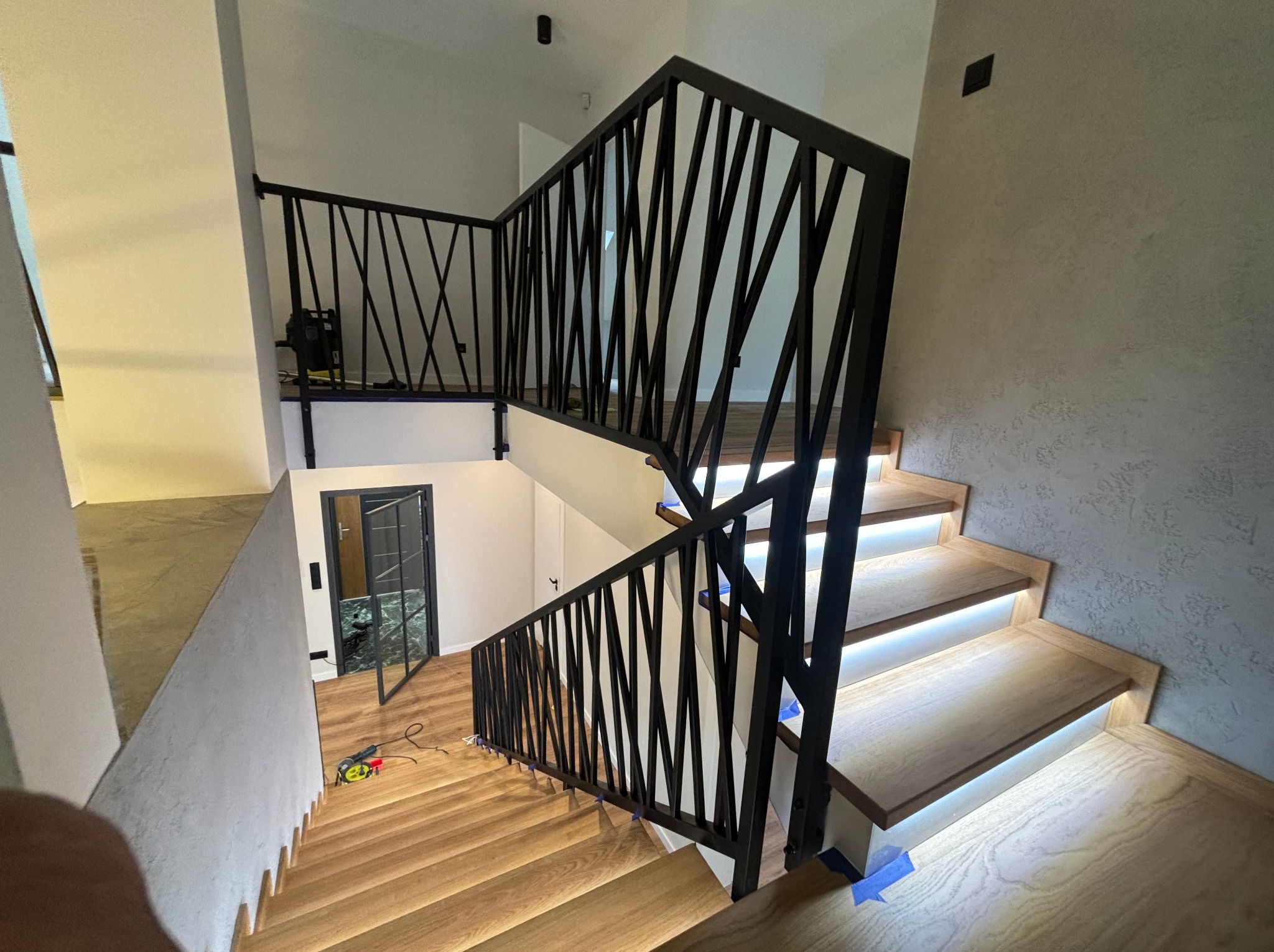 Nowoczesne schody z podświetlanymi stopniami i czarną, geometryczną balustradą. Widok z góry na dół, z narzędziami widocznymi na podeście.