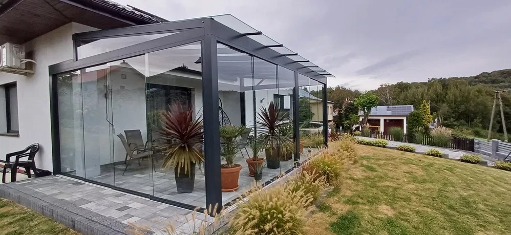 Szklana zabudowa tarasu z przesuwnymi panelami, czarna aluminiowa konstrukcja, rośliny w donicach, widok na ogród w tle.