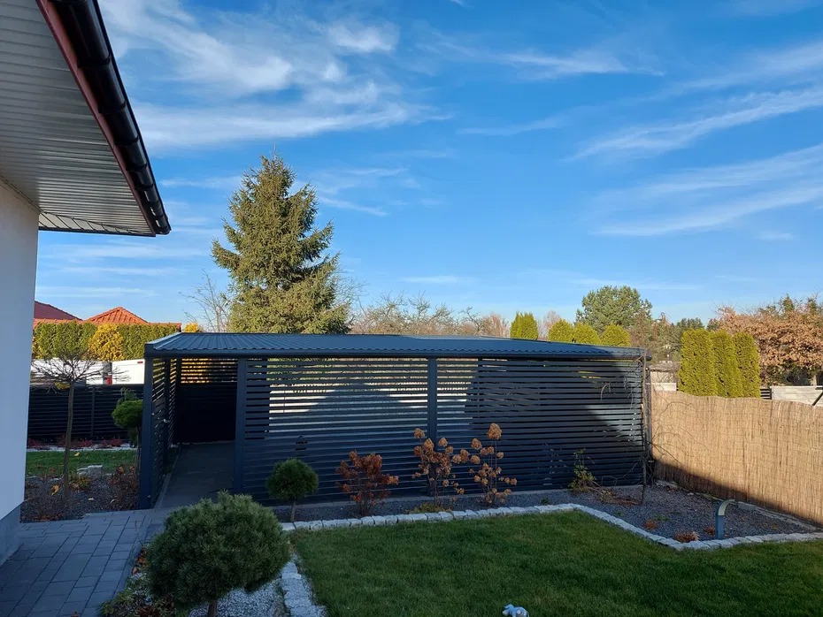 Nowoczesna, pozioma, metalowa konstrukcja osłonowa w ogrodzie, z widocznymi detalami wykończenia i otaczającą zielenią, w słoneczny dzień.