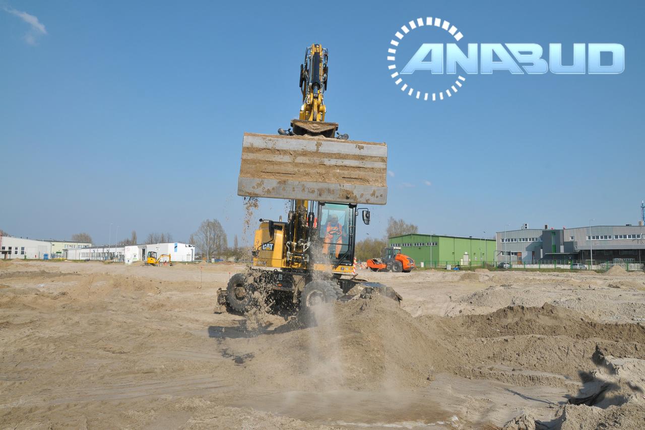 Żółta koparka kołowa CAT wysypuje ziemię na placu budowy, w tle budynki i walec, logo firmy ANABUD na niebie.