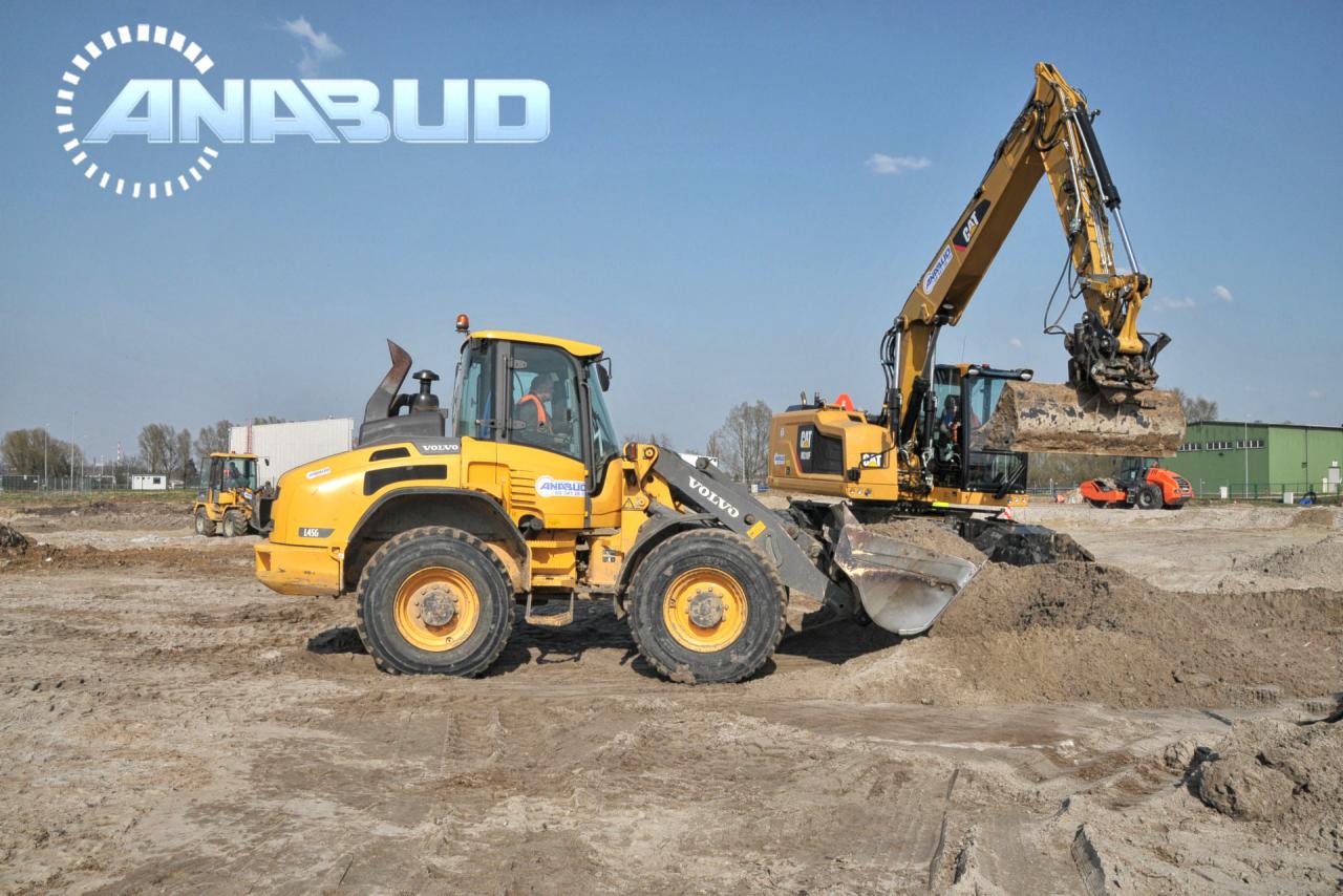 Żółta ładowarka Volvo i koparka CAT pracują na placu budowy, w tle widoczny budynek przemysłowy i inne maszyny budowlane, logo firmy ANABUD na niebie.