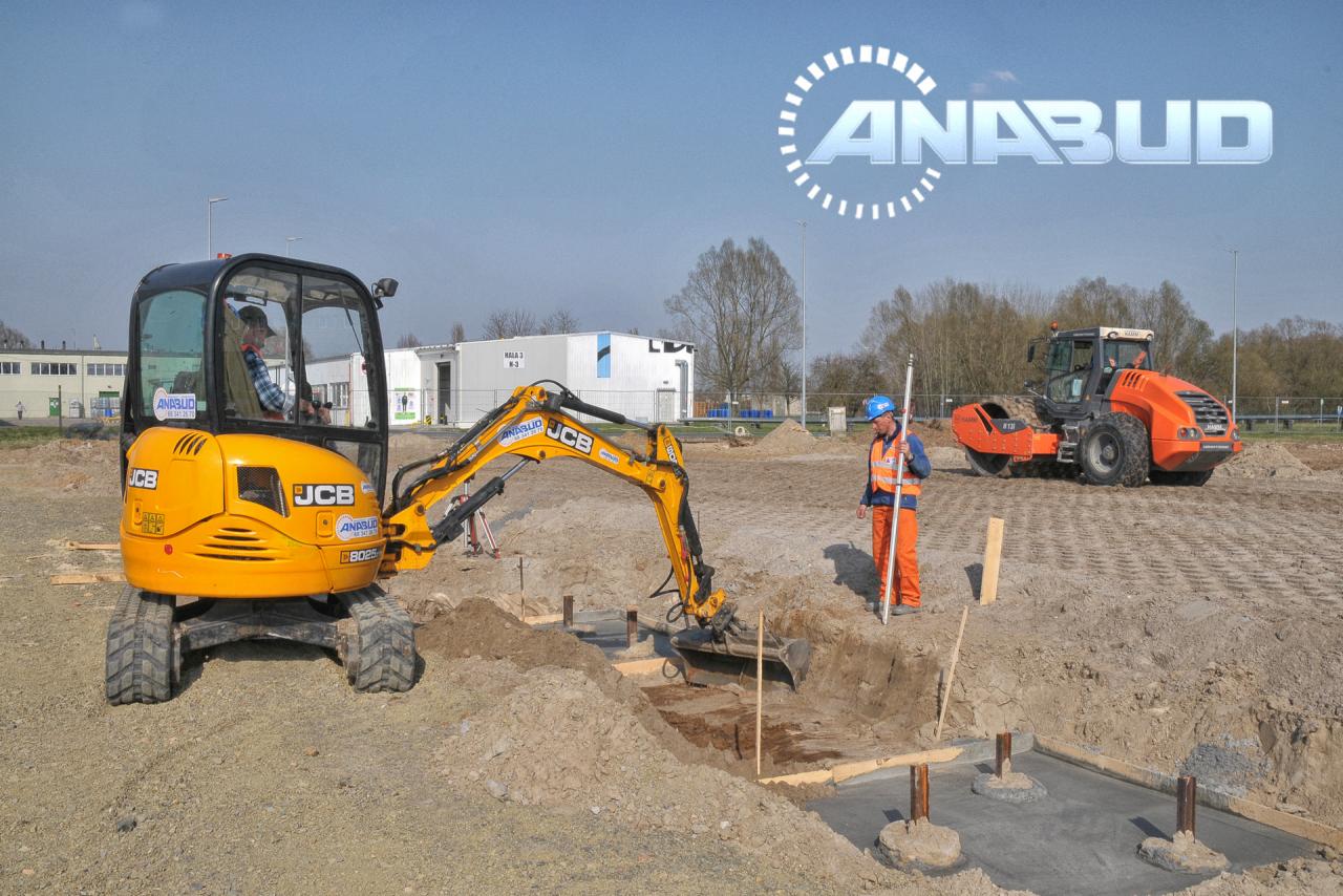 Żółta mini koparka JCB przygotowuje wykop pod fundamenty, obok stoi pracownik w pomarańczowym kombinezonie z łatą mierniczą, w tle walec i budynek.