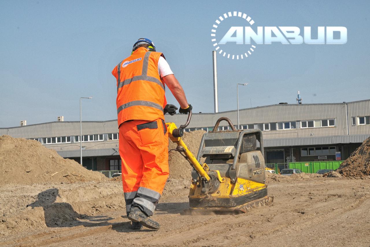 Pracownik w pomarańczowym kombinezonie zagęszcza grunt wibracyjną zagęszczarką Wacker, w tle hałda ziemi i budynek przemysłowy z logo firmy ANABUD.
