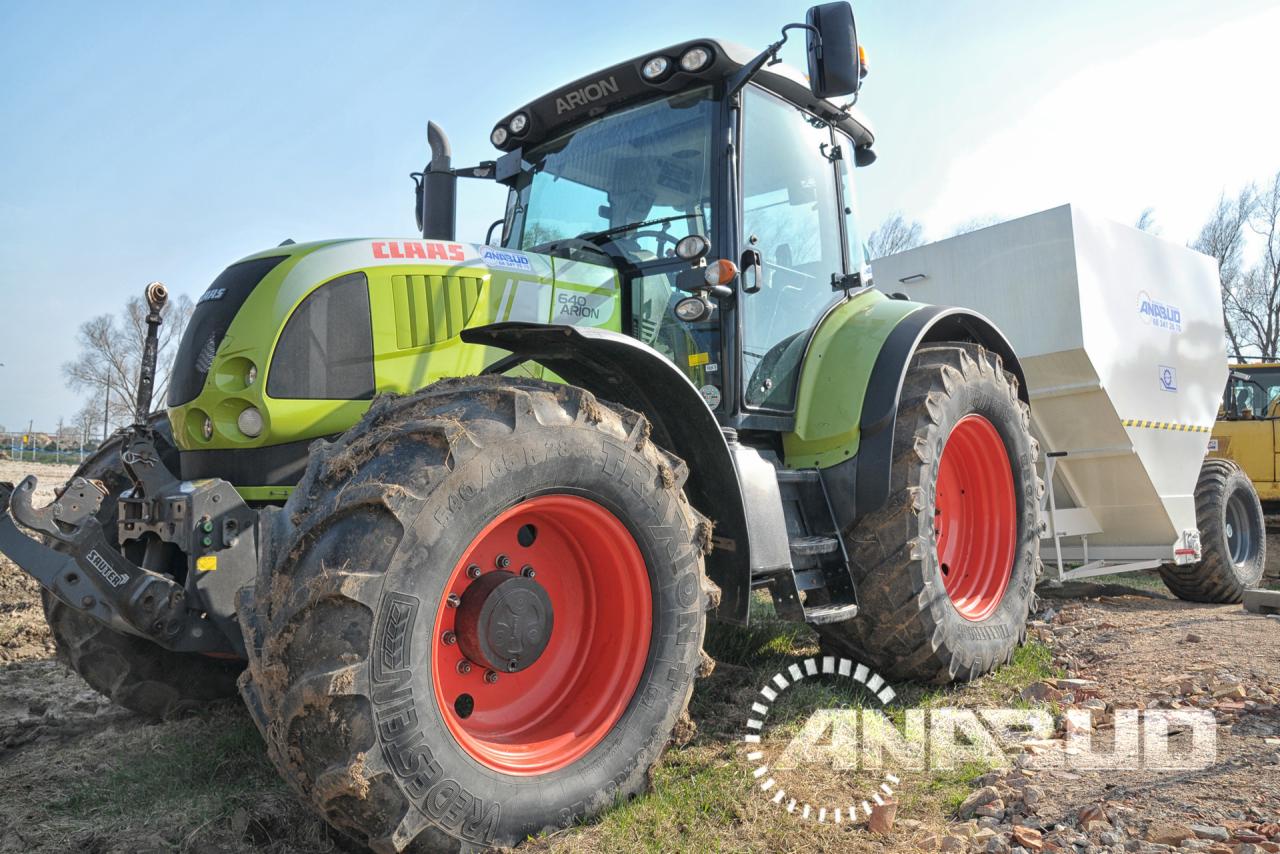 Zielono-żółty traktor Claas Arion z czerwonymi felgami i białym zbiornikiem na przyczepie, stojący na ziemi z elementami trawy, widok z niskiej perspektywy.
