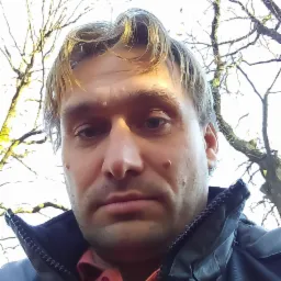 Portret mężczyzny z bliska, w kurtce i koszuli, z drzewami w tle. Ujęcie z dołu, podkreślające rysy twarzy i fakturę skóry.