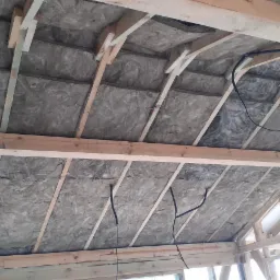 Izolacja z wełny mineralnej zamontowana między krokwiami dachu w konstrukcji drewnianej domu w trakcie budowy, widoczne przewody elektryczne.
