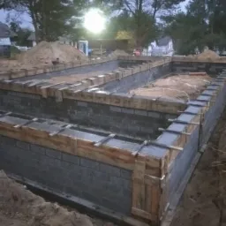 Wylane fundamenty z szalunkiem i bloczków betonowych na placu budowy domu jednorodzinnego, widoczny stos piasku, w tle drzewa i budynki.