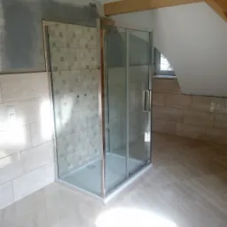 Montaż kabiny prysznicowej z przesuwnymi drzwiami w łazience na poddaszu, widoczne beżowe płytki na ścianach i podłodze, ściana częściowo w stanie surowym przed położeniem płytek, drewniana belka...