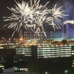 Nocne fajerwerki rozświetlające panoramę miasta z widocznymi budynkami biurowymi i w oddali wzgórzami.