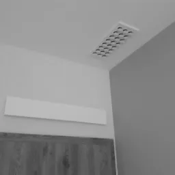 Nawiewnik sufitowy z okrągłymi otworami wentylacyjnymi, zamontowany w jasnym pomieszczeniu z dekoracyjną tapetą i widoczną górną częścią drzwi.