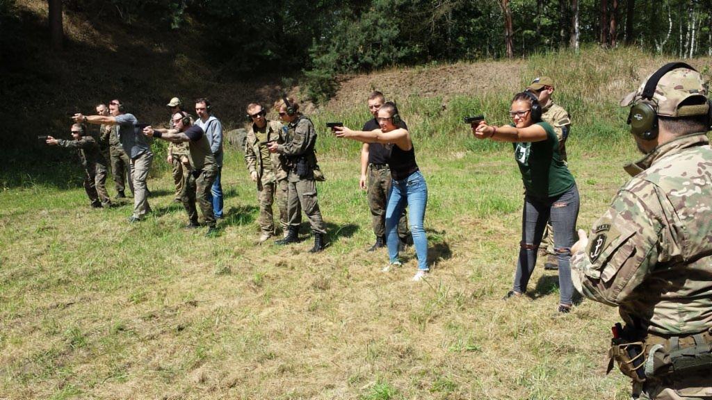 Grupa osób, w tym kobiety i mężczyźni, w różnym wieku, uczestniczy w treningu strzeleckim na otwartym terenie trawiastym, z drzewami w tle. Niektórzy ubrani w mundury wojskowe, inni w ubrania...