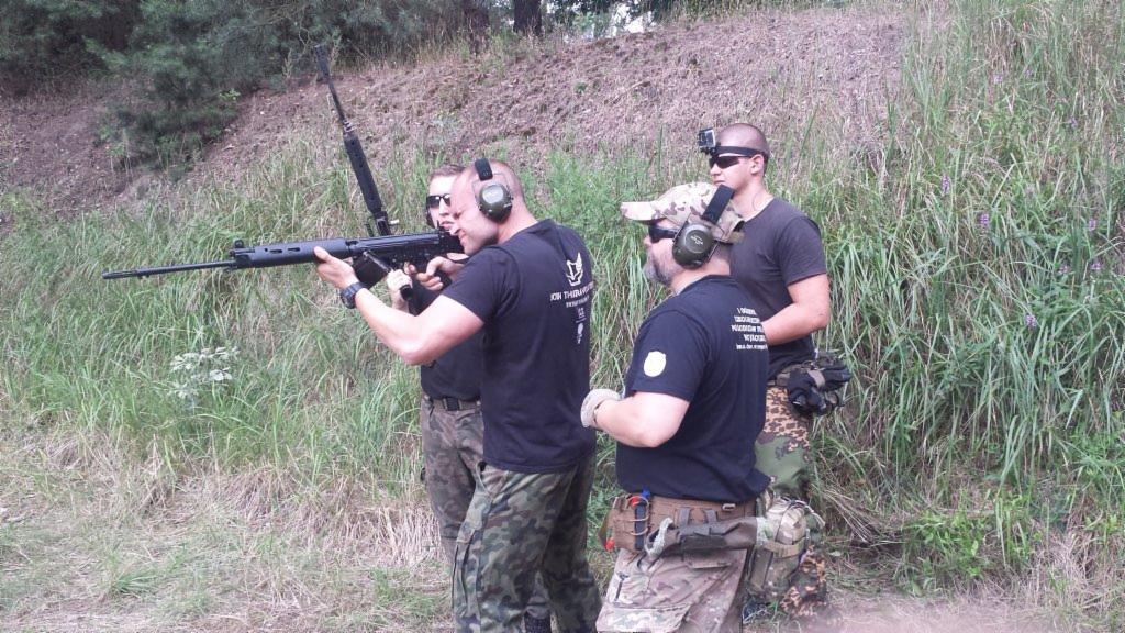 Mężczyźni w kamuflażu z bronią na tle zarośli, prawdopodobnie podczas treningu strzeleckiego.
